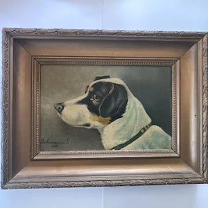May include: An antique framed painting of a dog in profile. The dog has a white coat with black markings on its head and ears. The painting is signed "Schuermans J. 1923" and is set in a decorative, ornate wooden frame.