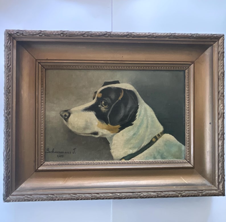 May include: An antique framed painting of a dog in profile. The dog has a white coat with black markings on its head and ears. The painting is signed "Schuermans J. 1923" and is set in a decorative, ornate wooden frame.
