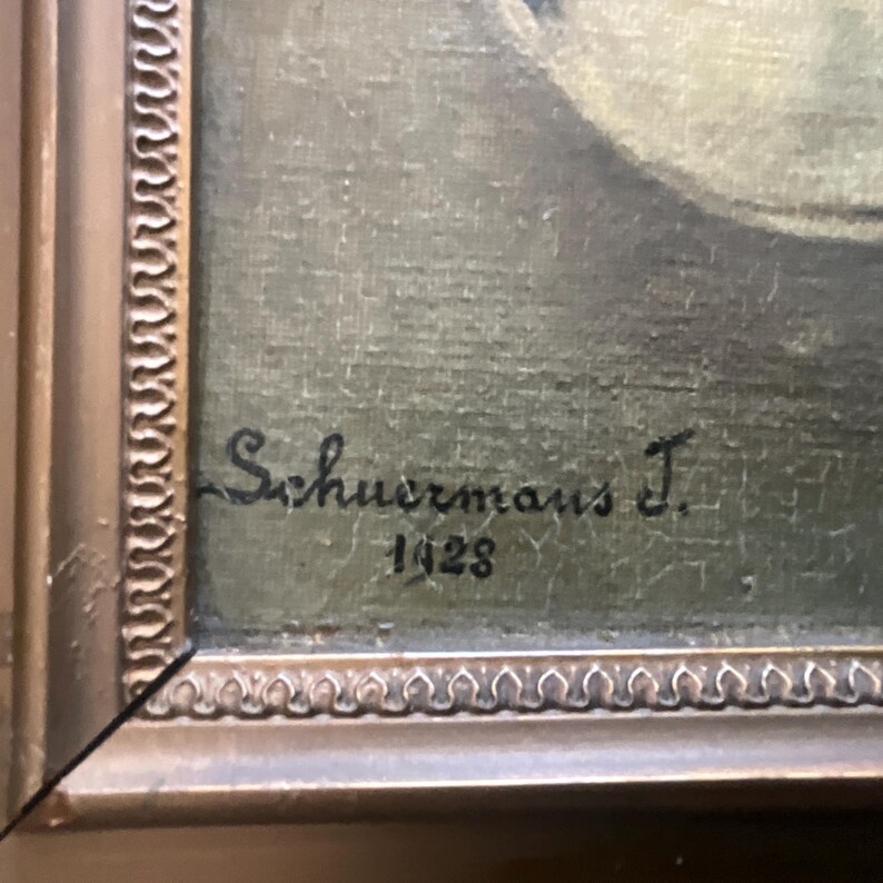 May include: Close-up of a framed painting with a gold-colored ornate frame. The painting features a signature that reads "Schuermans J." and the year "1928" in black ink. The artwork has a muted color palette.