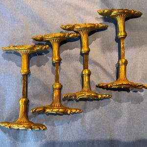 May include: Four gold-coloured metal door handles with ornate, leaf-like decorative plates at each end. Each handle features a central fluted column and a square-shaped spindle connecting the two ends, showcasing a metallic finish and detailed relief.
