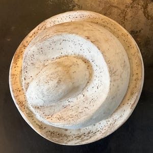 Könnte beinhalten: Ein kreisförmiges, cremefarbenes Dekorationsobjekt mit einem Spiraldesign. Das Objekt hat eine strukturierte Oberfläche mit dunklen Sprenkeln und einem erhabenen, geschichteten Aussehen. Der äußere Rand hat ein etwas dunkleres, gealtertes Aussehen.