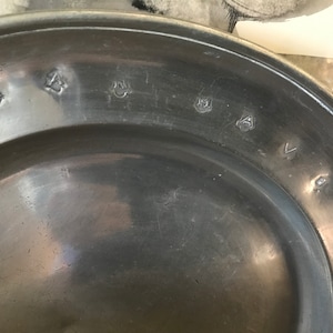 May include: Close-up of the bottom of a pewter plate with several markings. The markings include a crown, a letter "M", and a number "4".