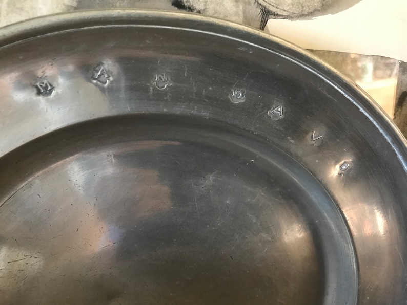 May include: Close-up of the bottom of a pewter plate with several markings. The markings include a crown, a letter "M", and a number "4".