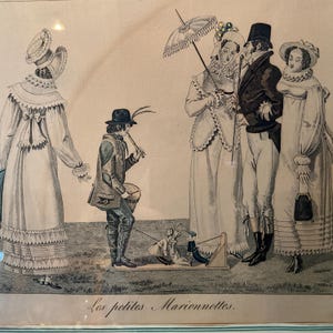 May include: An antique print depicting a scene with figures in period clothing. A man plays a flute while marionettes are displayed on a small stage. Several elegantly dressed people stand nearby. The print is framed and includes the text "Les petites Marionnettes."