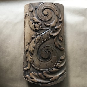Op de afbeelding: Een houten carving met een gedetailleerd ontwerp van draaiende bladeren en wijnstokken. De carving is donkerbruin van kleur en heeft een gladde, gepolijste afwerking.