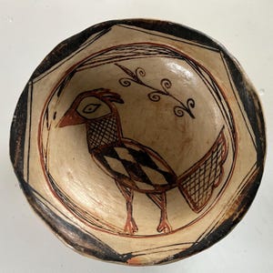 May include: A round ceramic bowl with a cream-coloured interior featuring a hand-painted black and reddish-brown bird illustration. The bird has a cross-hatched neck and tail, with a geometric body pattern. The bowl rim has a thick black border.