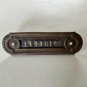 Puede incluir: Una placa de buzón rectangular de color bronce con la palabra "LETTRES" en relieve en el centro. La placa tiene detalles decorativos en los bordes y dos agujeros circulares para el montaje.