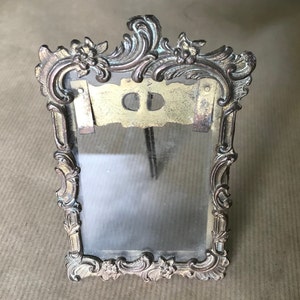 May include: An ornate silver-toned picture frame with a glass insert. The frame features a decorative design with floral motifs.