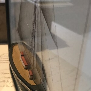 May include: A small wooden model of a sailboat with a white sail and a black hull. The model is framed and displayed on a wall.