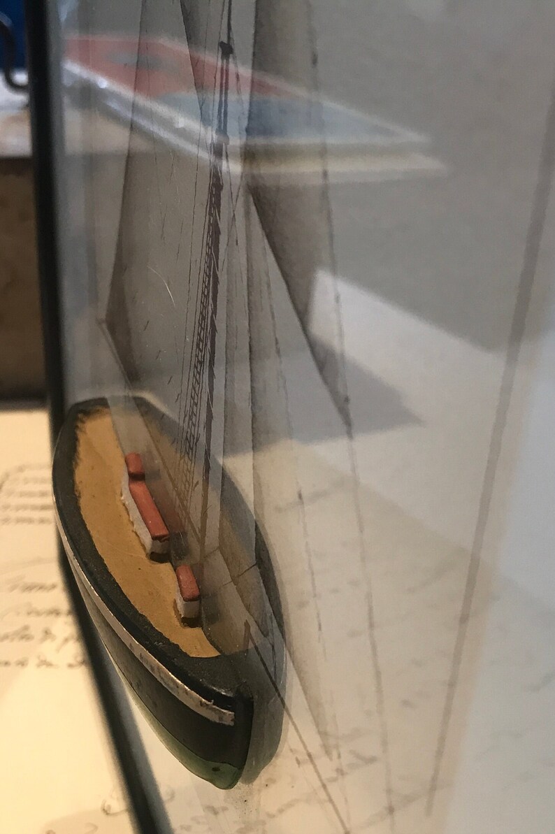 May include: A small wooden model of a sailboat with a white sail and a black hull. The model is framed and displayed on a wall.