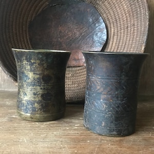 Puede incluir: Dos tazas antiguas de latón con un aspecto envejecido y desgastado. Las tazas tienen forma cilíndrica con un borde ligeramente ensanchado. Una taza tiene bandas horizontales, mientras que la otra tiene un diseño estampado. El fondo incluye una cesta tejida y una superficie de madera.