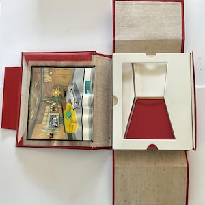 May include: An open red box with a white interior and a pop-up diorama. The diorama shows a room with a yellow sofa, a computer, and a window. The box has a red exterior and a beige interior.