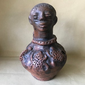 Peut inclure: Une sculpture en céramique de terre cuite représentant la tête et le torse d'une personne. La sculpture est recouverte d'un glacis brun et présente un visage avec un large sourire. Le torse est décoré d'un motif texturé et de trous.