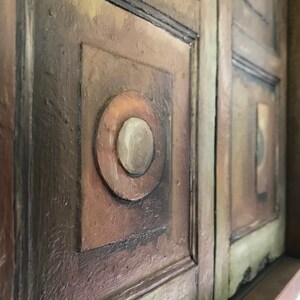 Op de afbeelding: Een close-up van een houten wandkunstwerk met een geometrisch ontwerp. Het stuk is geschilderd in tinten bruin, groen en wit. Het ontwerp bevat een reeks vierkanten en cirkels.