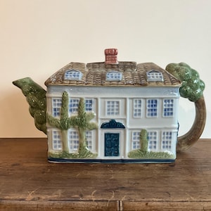 Peut inclure: Une théière en céramique blanche en forme de maison avec un toit brun, une cheminée rouge et des arbres verts sur les côtés. La théière a des fenêtres bleues et une porte bleu foncé.