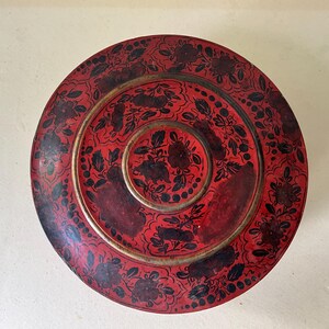 Può includere: Una scatola rotonda in lacca rossa con un motivo floreale nero dipinto a mano sul coperchio. Il design presenta un motivo circolare centrale circondato da un anello di fiori e foglie, con un bordo color metallo che separa le sezioni.
