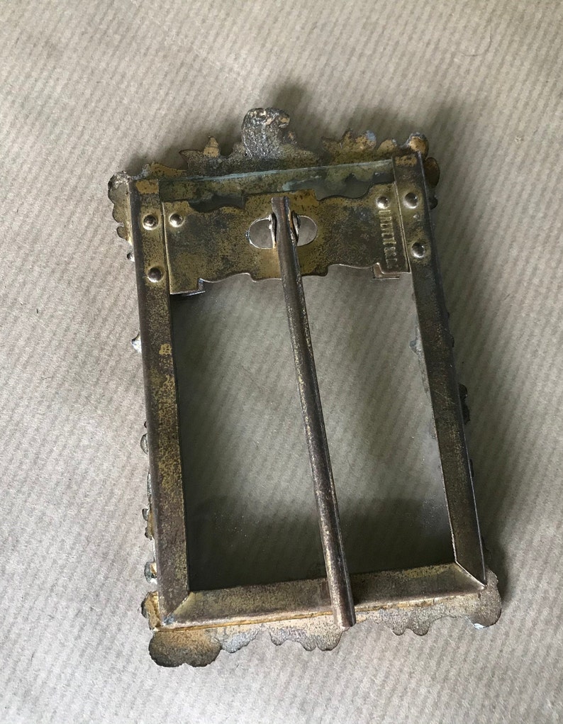 May include: A gold-toned metal picture frame with an ornate design. The frame has a glass front and a metal stand on the back. The back of the frame has the inscription "W.H. MILLER & CO."