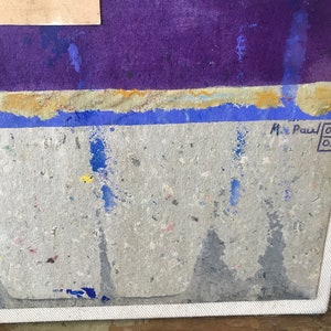 May include: A close-up of a textured, grey canvas with blue and yellow paint splatters. The canvas is framed in white and has a signature in the bottom right corner that reads "M. Paul 06 02".