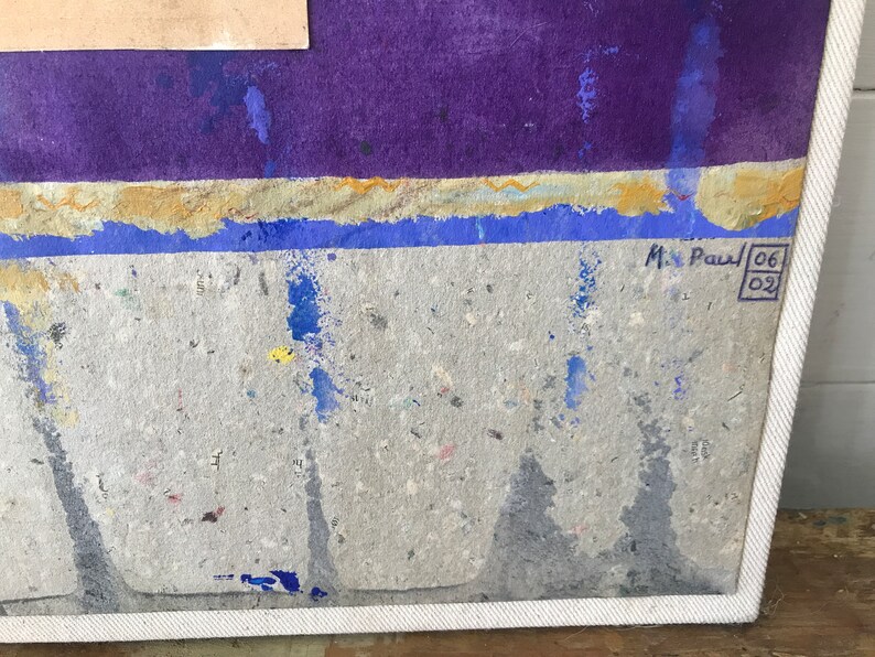 May include: A close-up of a textured, grey canvas with blue and yellow paint splatters. The canvas is framed in white and has a signature in the bottom right corner that reads "M. Paul 06 02".