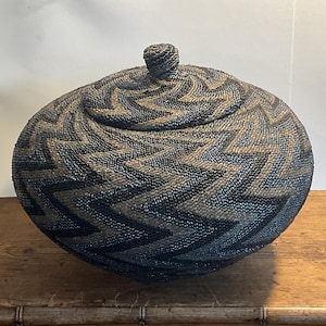 May include: A large, round, black and brown woven basket with a lid. The basket is made of small, tightly woven beads and has a geometric pattern.