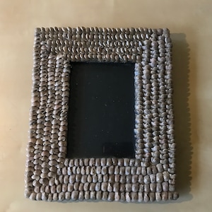 Peut inclure: Un cadre photo rectangulaire avec une bordure faite de petits galets de couleur claire. Le cadre a un intérieur sombre et noir, et les galets sont disposés en rangées soignées. Le cadre est de couleur neutre et complèterait divers styles de décoration.