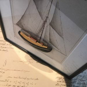 May include: A framed black and white drawing of a sailboat with a small wooden model of a sailboat in the centre of the frame. The drawing is on a white background and the model is made of wood and painted brown.