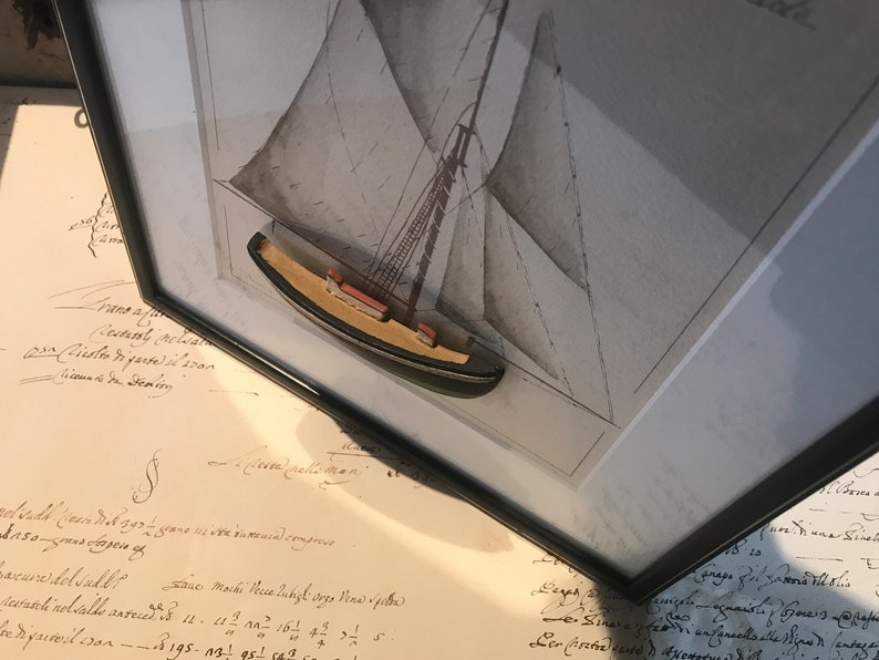 May include: A framed black and white drawing of a sailboat with a small wooden model of a sailboat in the centre of the frame. The drawing is on a white background and the model is made of wood and painted brown.
