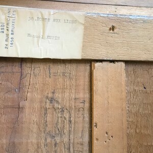 Op de afbeelding: Een close-up van een houten frame met een label eraan bevestigd. Het label luidt "EL AMANECER EQUATE asbl 24 RUE AFRICAINE 1050 BRUXELLES 38-PORTE AUX LICONS".