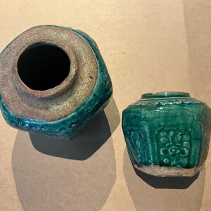 Puede incluir: Dos tarros de cerámica hexagonales con un esmalte verde vibrante. El tarro más grande se muestra desde una perspectiva cenital, revelando una amplia abertura circular y un borde rugoso sin esmaltar. El tarro pequeño tiene motivos florales.