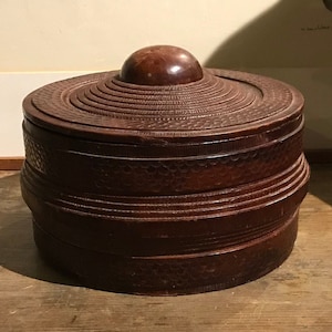 May include: A round, wooden box with a carved lid and a rounded knob on top. The box is dark brown and has a layered, concentric design.