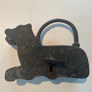 May include: A dark brown cast iron padlock shaped like a lion. The lock features a curved handle and a keyhole. The lion is depicted lying down. The design is rustic and the metal has a textured finish.