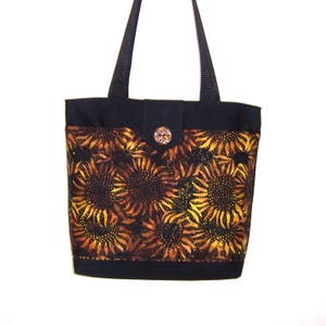 May include: A black tote bag with a sunflower pattern. The bag features a black top and handles, with a decorative button closure. The sunflower design is in shades of yellow, orange, and brown.