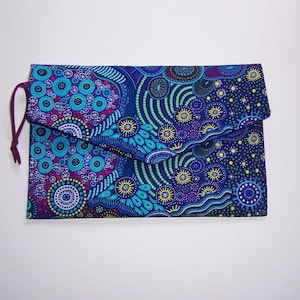May include: A rectangular clutch with a flap closure, featuring a vibrant, colorful pattern. The design incorporates circular and dot motifs in shades of blue, teal, purple, and yellow. A dark purple cord secures the flap. The clutch is likely made of fabric.