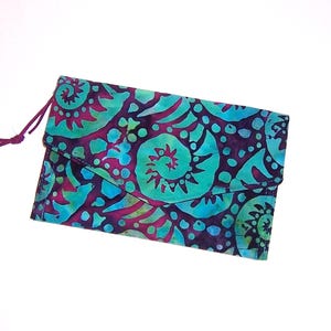 May include: A rectangular fabric clutch with a flap closure featuring a vibrant batik pattern in shades of teal, turquoise, and deep purple. The design displays swirling spiral motifs and circular dots throughout. A thin purple cord is attached to the side.