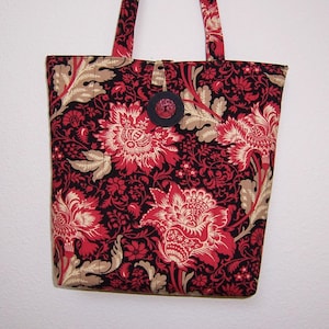 May include: A tote bag with a floral pattern in red, beige, and black. The bag has a button closure and matching handles. The fabric features large stylized flowers and foliage. The bag is designed for carrying items.