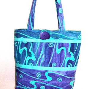May include: A handmade tote bag with a vibrant, abstract design. The bag features a swirling pattern in shades of purple and teal, with a teal band and a purple button closure. The handles and the body of the bag are made of the same fabric.