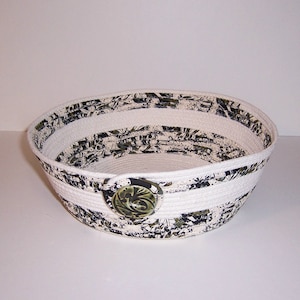 May include: A white and black fabric-covered bowl with a white rope trim and a green and black button on the side.