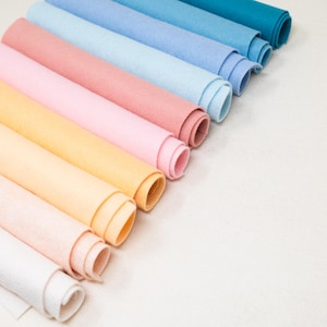 May include: A collection of rolled felt sheets in various pastel colors, including pink, peach, yellow, and blue. The felt rolls are arranged diagonally, showcasing the different shades and textures of the fabric. The background is a plain white surface.