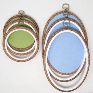 May include: A collection of oval embroidery hoops in various sizes. The hoops have a wood-grain finish with white inner rings and bronze-colored hardware. The fabric inside the hoops is light green and light blue.
