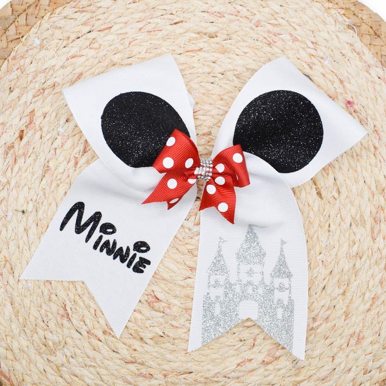 May include: A white hair bow with black glitter circles and a red polka dot bow. The word "Minnie" is printed in black script. A silver glitter castle is also featured on the bow.