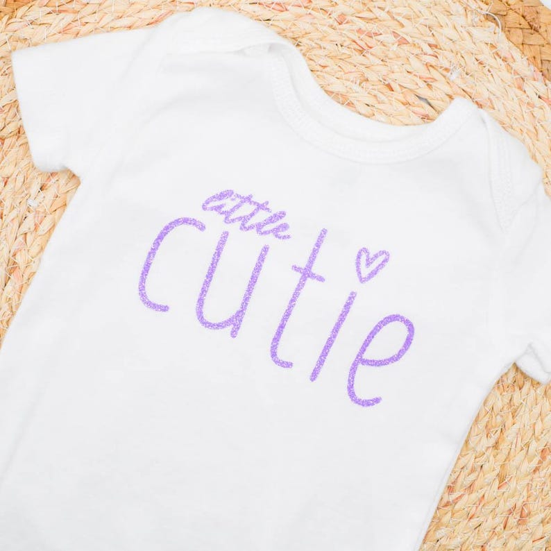 May include: White baby bodysuit with short sleeves. The bodysuit features the words "little cutie" in glittery purple script, with a small heart symbol. The garment is on a woven, light brown surface.