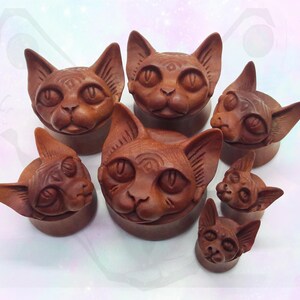 May include: Set of eight wooden cat-shaped ear gauges in various sizes. The gauges are carved with detailed features, including eyes, nose, and whiskers. The wood is a rich brown color.