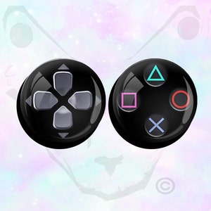 以下が含まれることがあります： PlayStationコントローラーのボタンに似た、黒く光沢のある2つの円形オブジェクト。1つは方向パッド、もう1つは緑の三角形、ピンクの四角、赤の円、青のXなどの色の付いたシンボルが付いています。背景はパステル調の銀河です。