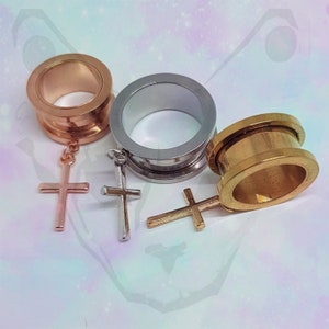May include: Three different coloured metal ear gauges with cross charms hanging from them. The gauges are silver, gold, and rose gold.