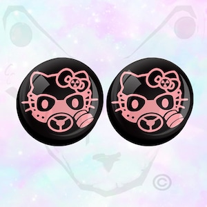 May include: Pair of black ear gauges featuring a pink Hello Kitty design wearing a gas mask. The design includes a bow, eyes, and a radiation symbol. The gauges are round and have a glossy finish.