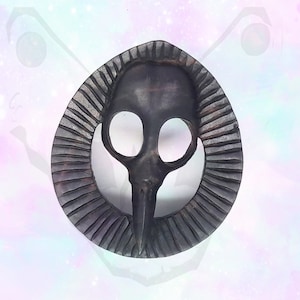 May include: A black metal pendant featuring a detailed skull design with a long beak. The skull is surrounded by a circular frame with a textured, ridged pattern.