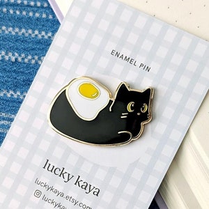 May include: Black enamel pin of a cat with a fried egg on its back. The pin is on a white card with the text "lucky kaya" and "lucky kaya.etsy.com" and "@lucky kaya.art". The card also says "enamel pin".