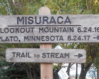 May include: A wooden sign with the words "Misuraca" and "Trail to Stream" carved into it. The sign also has directions to Lookout Mountain and Plato, Minnesota with dates 6.24.16 and 6.24.17.