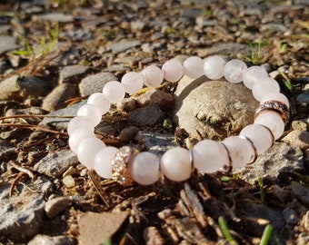 May include: A pink rose quartz beaded bracelet with a rose gold charm and clear rhinestones. The bracelet is lying on a bed of small rocks.