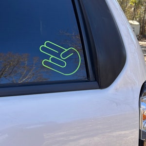 May include: A white car with a green hand gesture sticker on the back window.